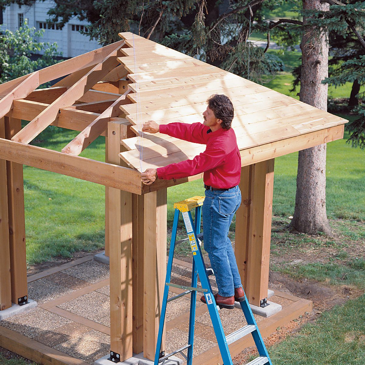 A person stands on a ladder while working on a wooden pergola, adjusting one of the rafters. The pergola's roof is partially assembled. The scene is set in a garden with green grass and tall trees in the background. The person wears a red shirt and blue jeans.