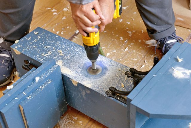 A persons hands are visible drilling holes for plumbing fixtures.