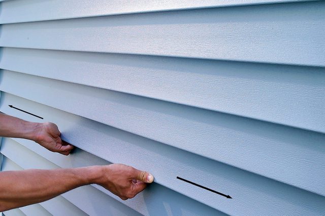 A person installs or adjusts horizontal vinyl siding panels on a building exterior. The image focuses on their hands placing a panel into position, showing the texture and alignment of several panels. The siding is light gray in color.
