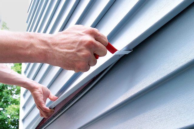 Close-up of a person using a utility knife to cut blue vinyl siding on the exterior wall of a house. The hands are holding the siding and the knife, with trees and a blurred background visible.