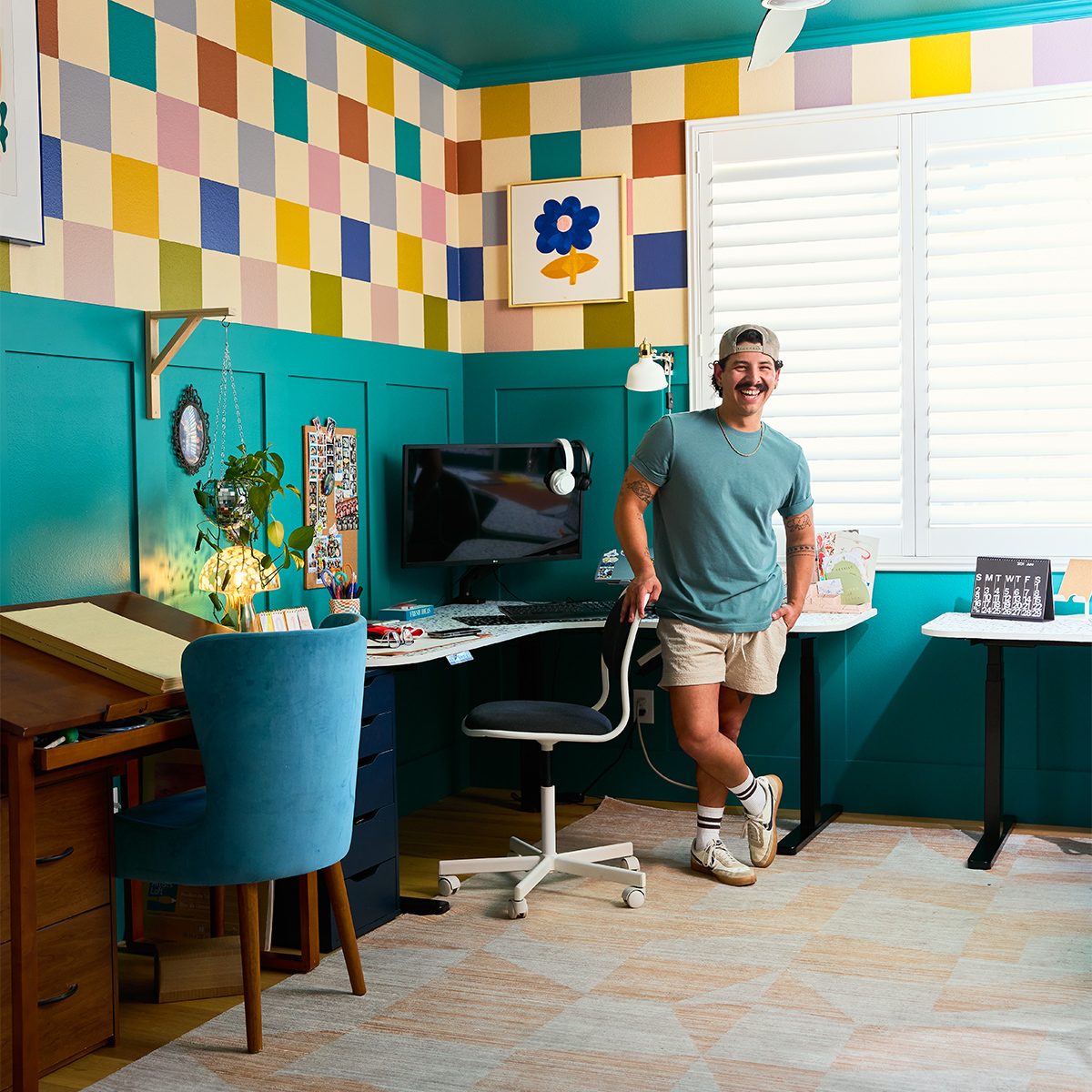 A person stands in a colorful, well-organized room featuring teal walls and a checkerboard pattern above. The room includes a desk with a computer, a drafting table, a chair with plush blue cushion, and various decorative elements, including a plant and artwork.