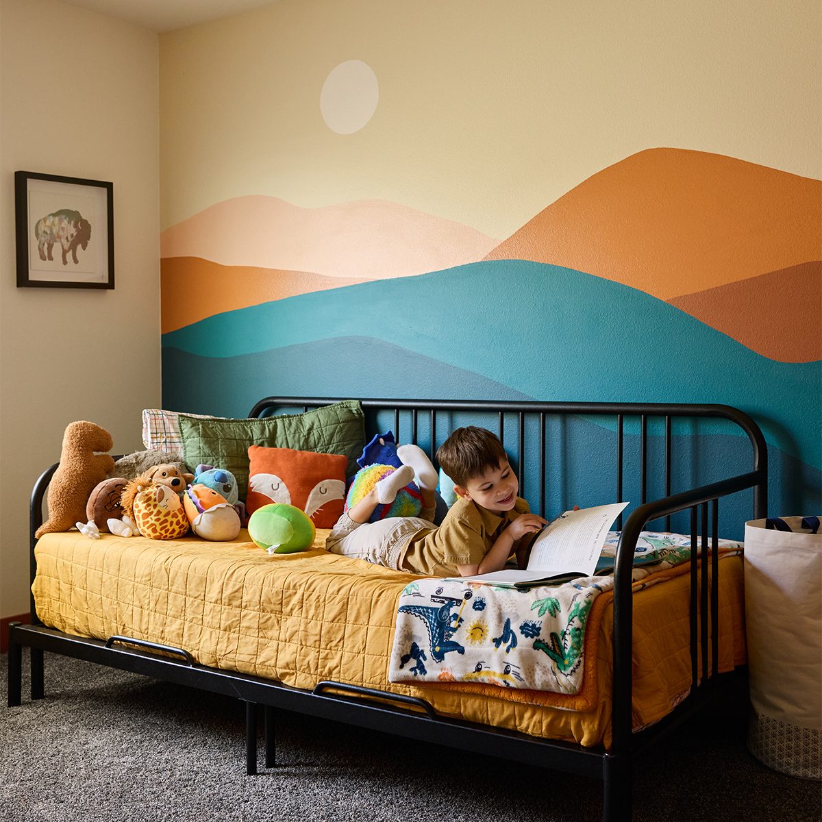 A young child lies on a bed with a yellow quilt, surrounded by plush toys. The wall behind the bed features a colorful mural of rolling hills in shades of blue, orange, and tan. The room also has a framed picture of an animal and a laundry basket.