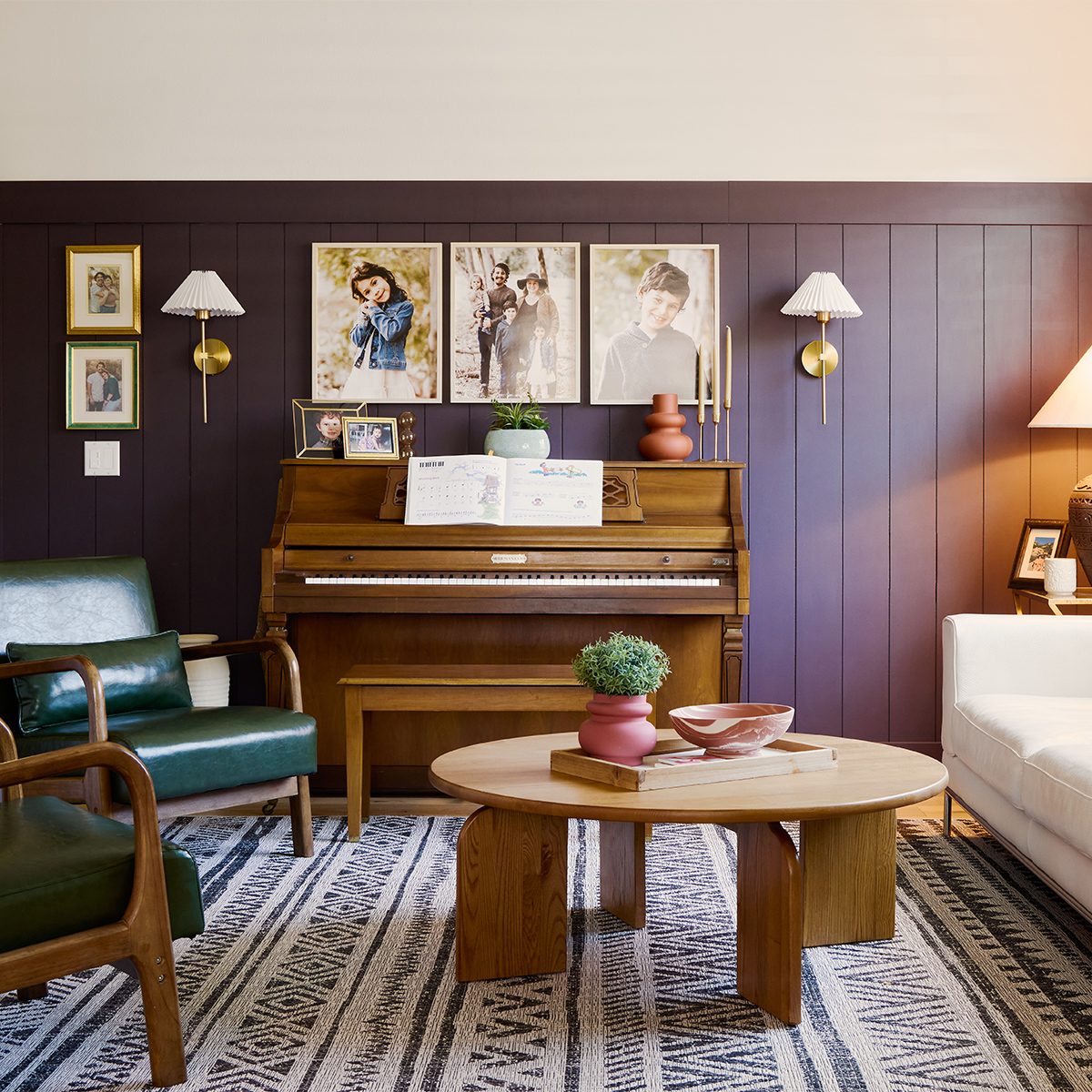 A cozy living room with a wood-paneled wall features a piano with sheet music, family photos, and sconces above. Green and white armchairs sit around a round wooden coffee table on a patterned rug. A table lamp and decorative items add a warm ambiance.