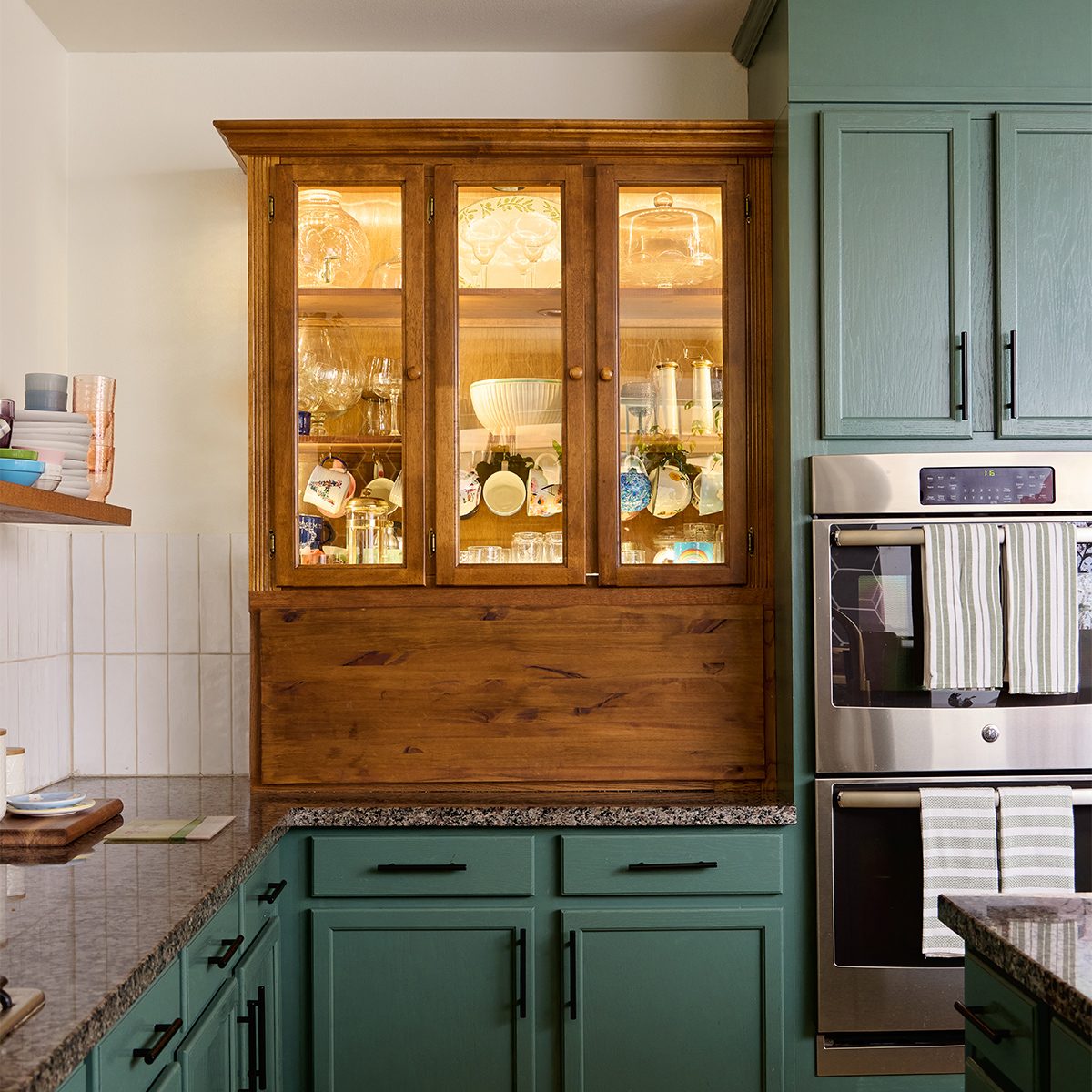 A kitchen with green cabinetry, granite countertops, and modern appliances. A wooden hutch with glass doors displays various dishes and glassware. A striped towel hangs from the oven handle. A shelf with books and a bowl is visible on the left wall.