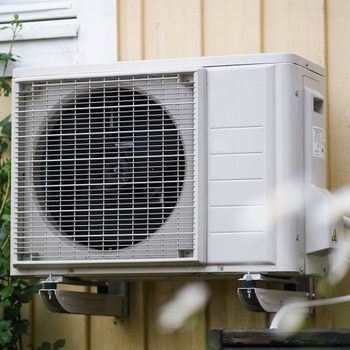 Outdoor HVAC unit mounted on the side of a yellow building. The unit has a large, circular fan with a protective grille on the front. Green foliage partially surrounds the bottom left corner of the unit.