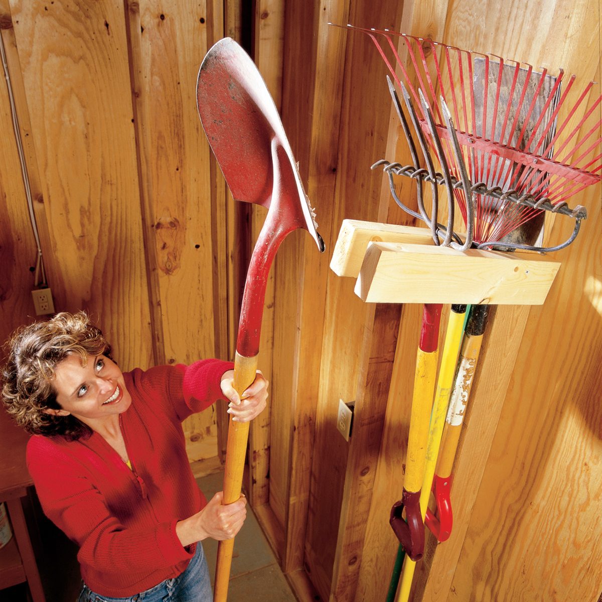 A person in a red shirt is holding a shovel and standing in a wooden shed. The shed is organized with gardening tools, including rakes and other long-handled implements, neatly hanging on a rack on the wall.