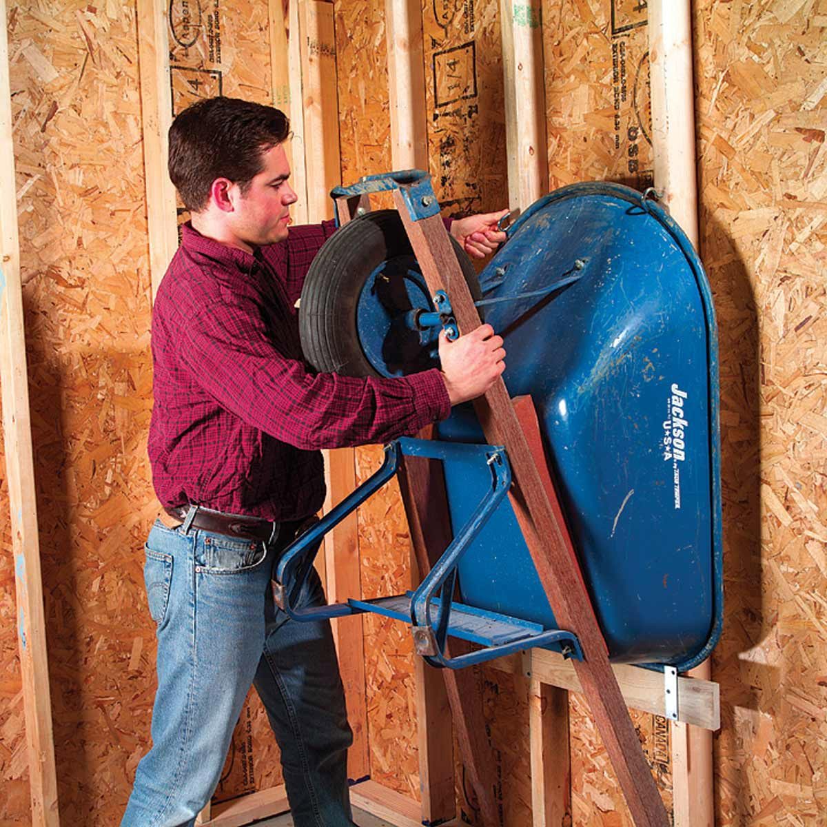 A person in a red shirt is lifting a blue wheelbarrow onto a wooden plank in a shed or an unfinished room with exposed plywood walls and wooden studs. The space appears to be under construction or being used for storage.