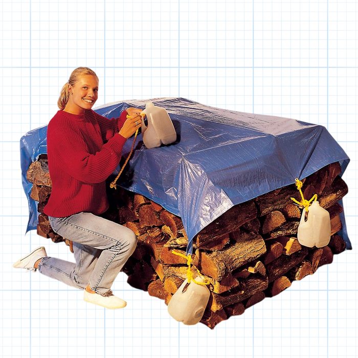 A person with light hair, wearing a red sweater and light blue jeans, is kneeling next to a stack of firewood. The firewood is covered with a blue tarp that is weighed down by white jugs tied to the tarp's corners.