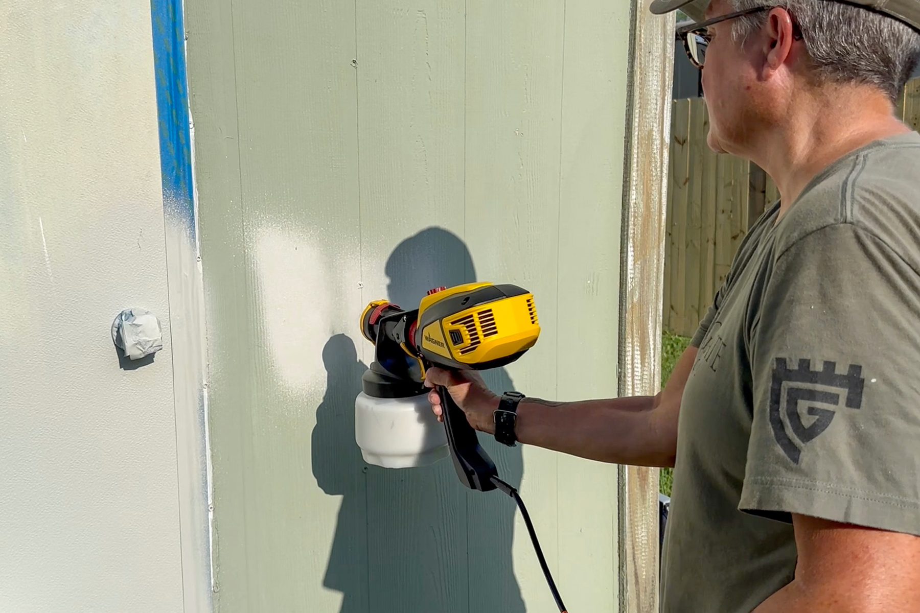 A person wearing a beige cap, gray t-shirt, and glasses uses a yellow and black paint sprayer to paint a green vertical surface. The person is standing outdoors next to a wooden structure.