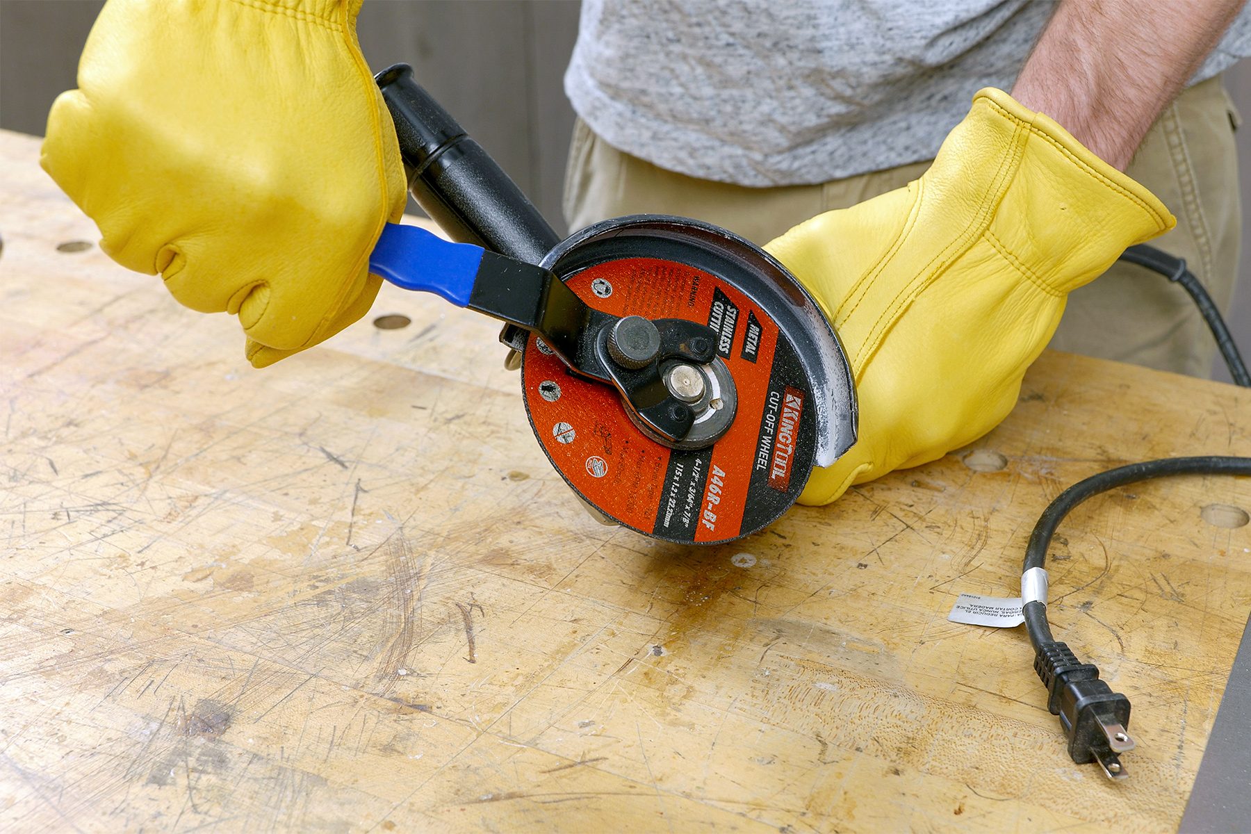 a close-up of a person preparing an angle grinder for use; The individual is wearing yellow protective gloves and appears to be adjusting or securing the cutting disc onto the angle grinder; The angle grinder is placed on a wooden workbench, The person is standing next to the workbench, ready to proceed with a metal cutting task;