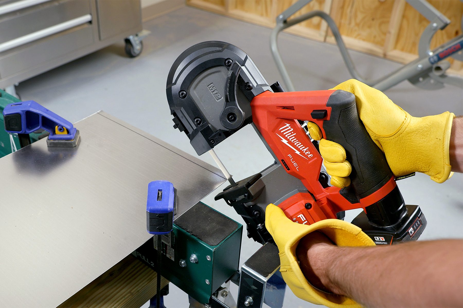 A person wearing yellow gloves uses a red Milwaukee-branded handheld power tool to cut through a metal sheet clamped to a workbench in a workshop. The background includes various tools and machinery.