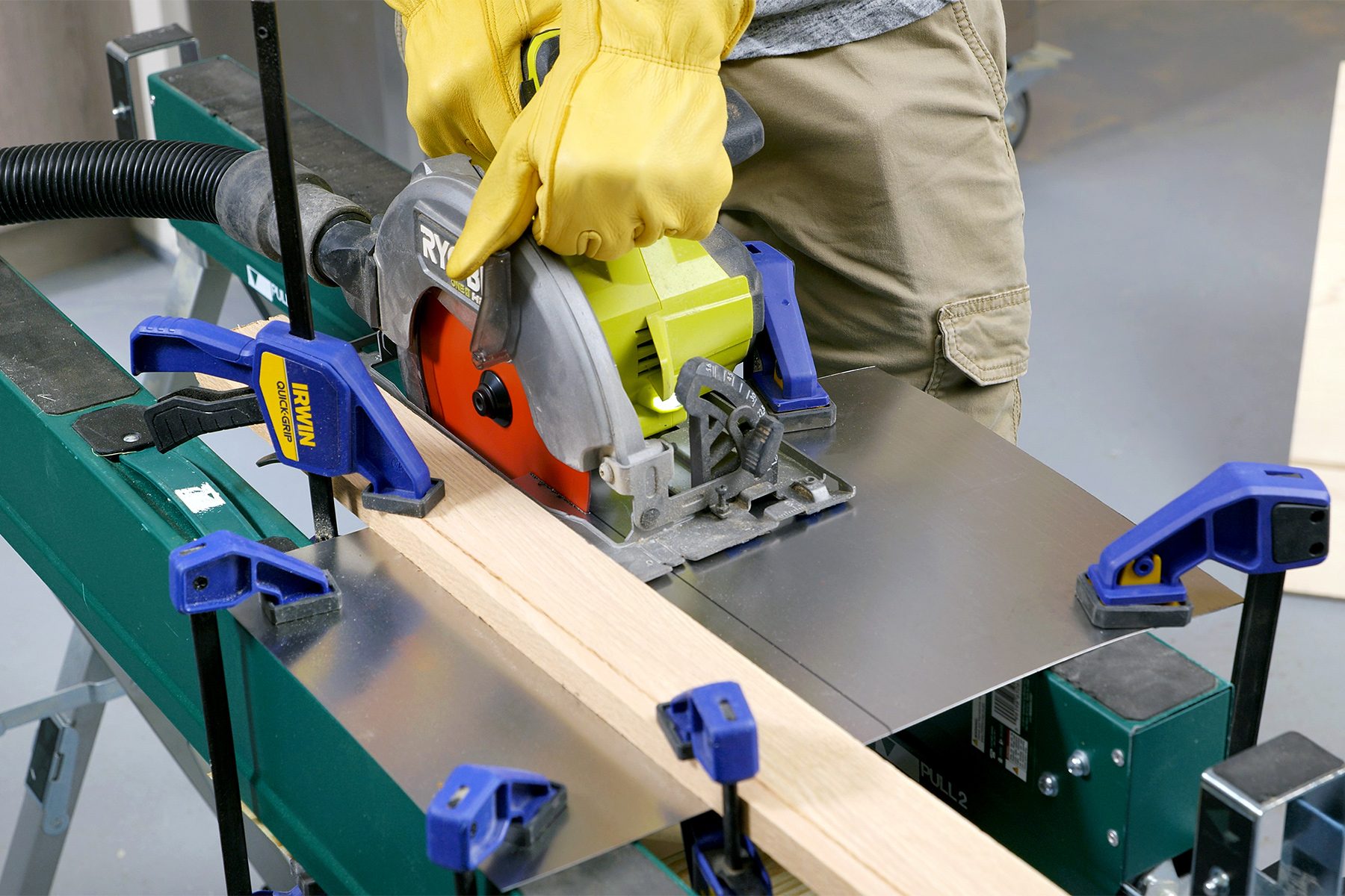 A person wearing yellow gloves is using a circular saw to cut a piece of wood supported by clamps on a workbench. Various clamps secure the wood in place, and the person stands over the bench, carefully guiding the saw along the wood.