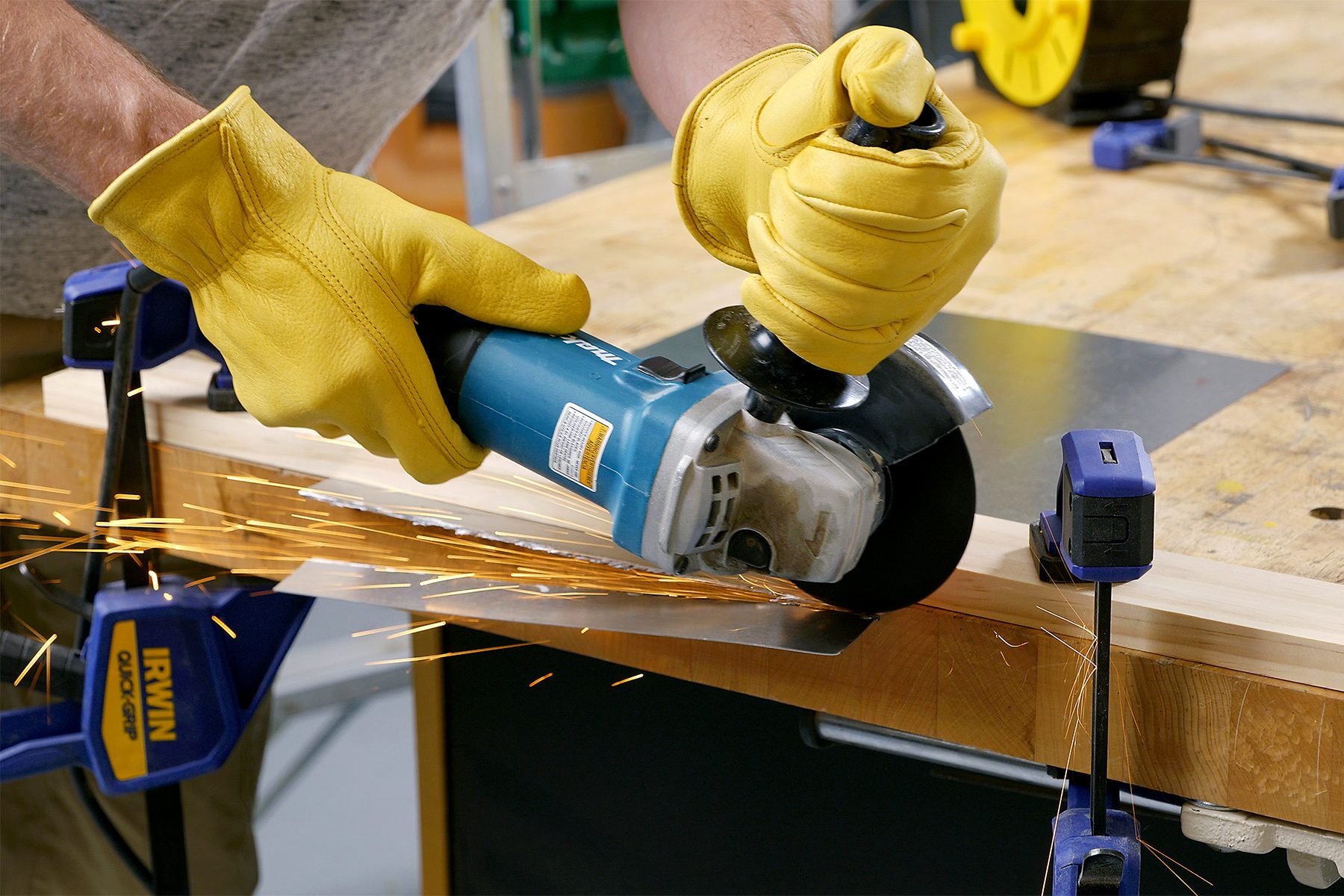 A person wearing yellow gloves is using a blue angle grinder to cut through a piece of metal that is clamped to a workbench. Sparks are flying from the metal as it is being cut. The workbench is equipped with blue clamps to hold the material in place.