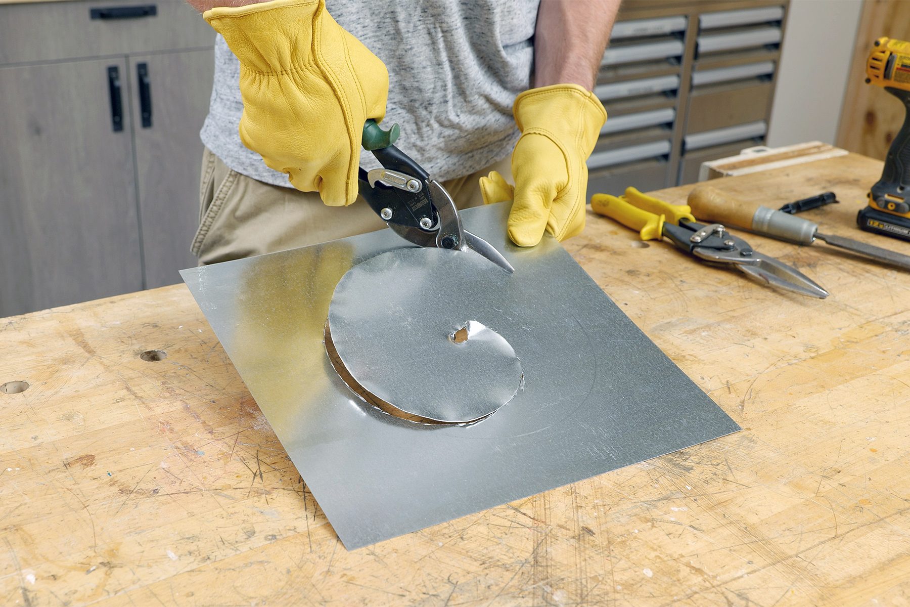 A person wearing yellow gloves is using tin snips to cut a circular shape out of a sheet of metal on a wooden workbench. Various tools are scattered on the bench, including another pair of snips, a drill, and tape measures.