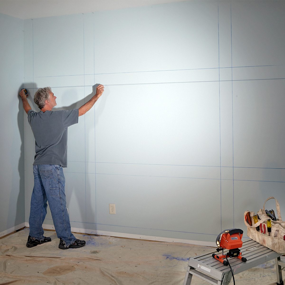 A person wearing a gray t-shirt and blue jeans is drawing horizontal lines on a light blue wall using a measuring tool. The floor is covered with a protective sheet, and there is a red power tool on a table nearby. The room appears to be under renovation.