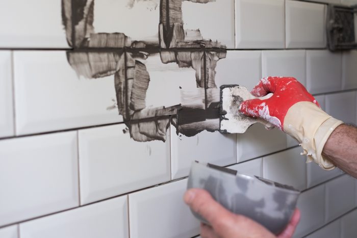 Worker applies grey grout at white tiles with rubber trowel.