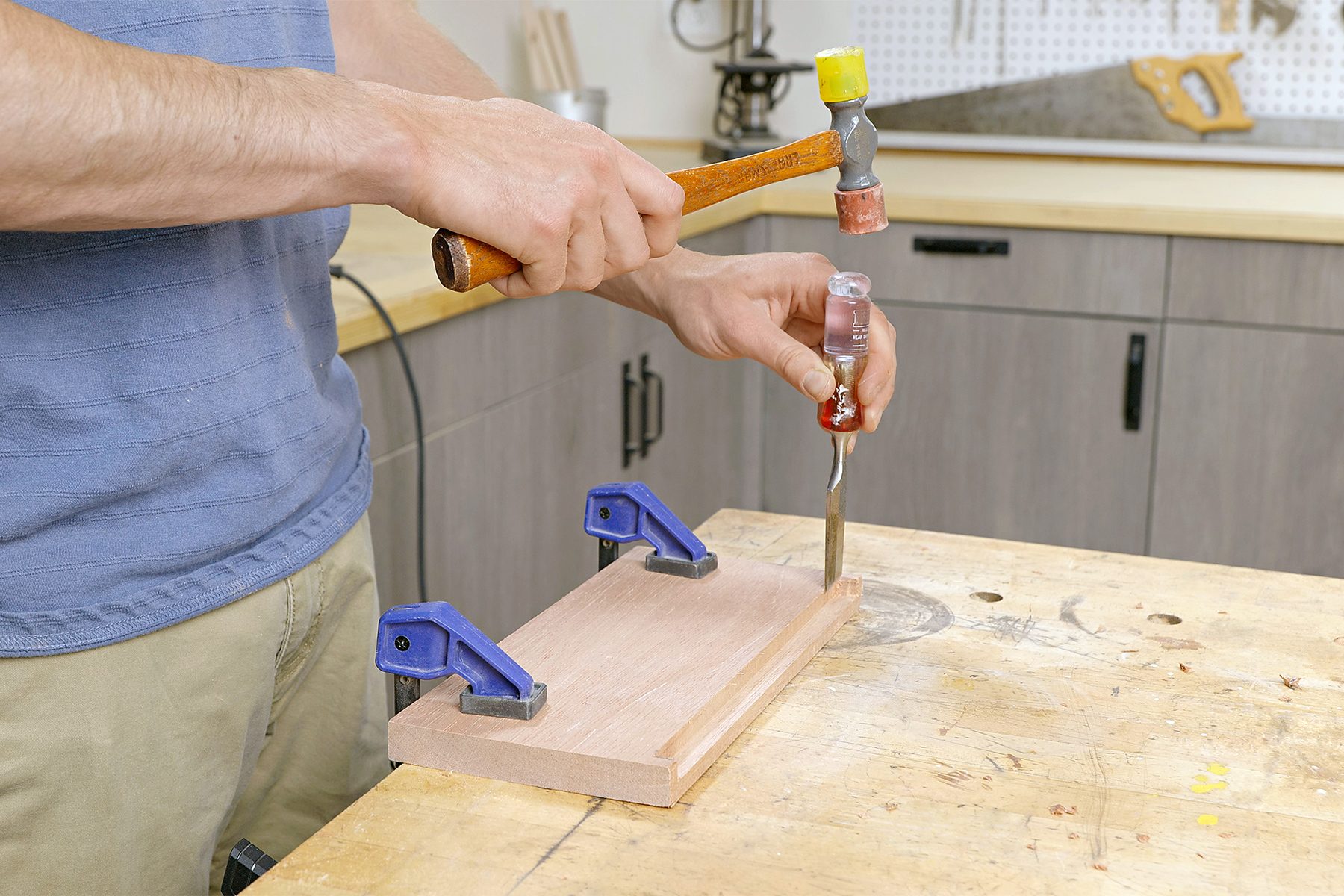 A person in a workshop taps a chisel with a hammer, working on a piece of wood secured with clamps on a workbench. Tools and cabinets are visible in the background.