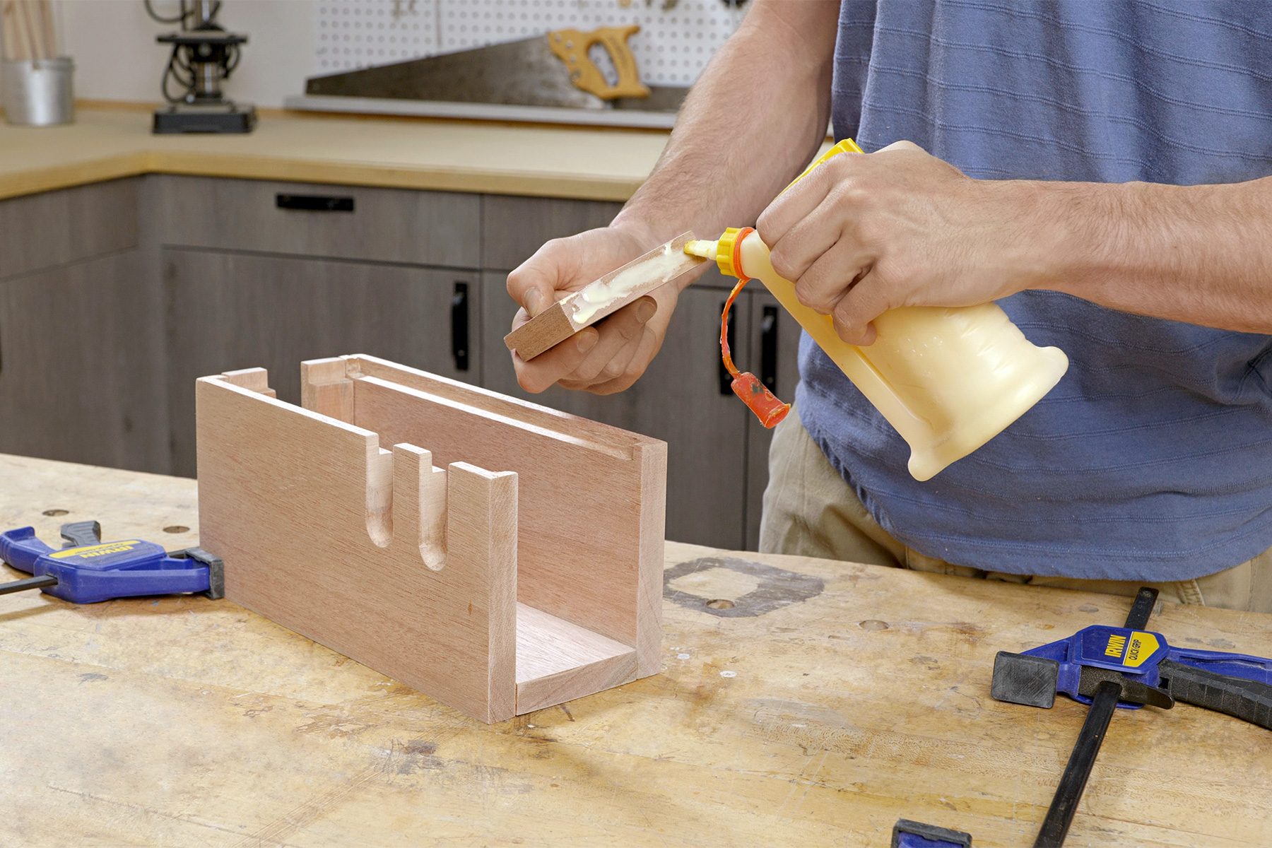 A person applies wood glue to a wooden joint using a bottle, assembling a rectangular wooden box. Clamps and other tools are visible on the workbench in a workshop setting.