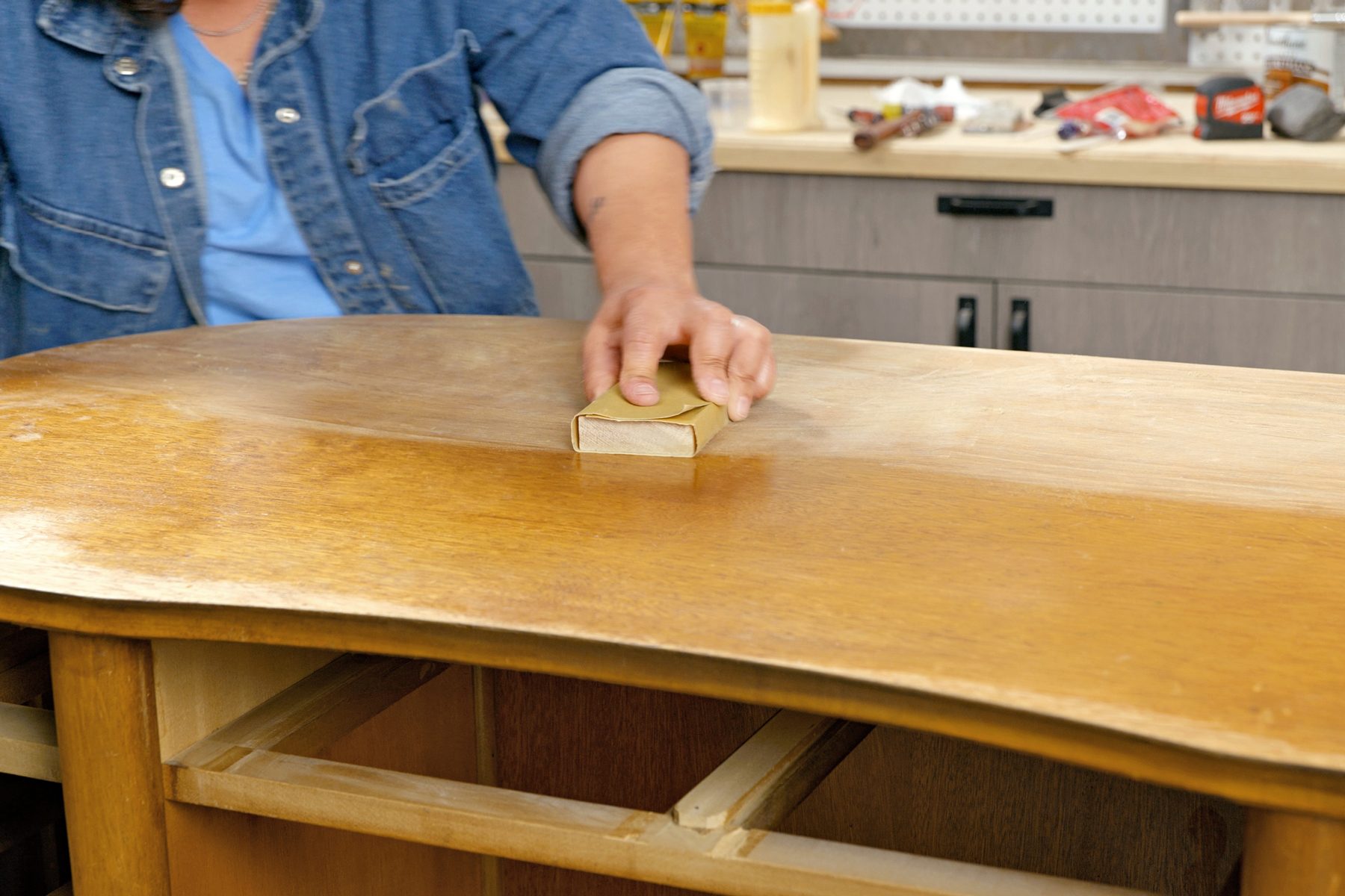 A man's hand is sanding a wooden surface with sandpaper; The wood is a light brown color, and the sandpaper is beige; The man is wearing a denim shirt; dust is visible on the surface of the wood;