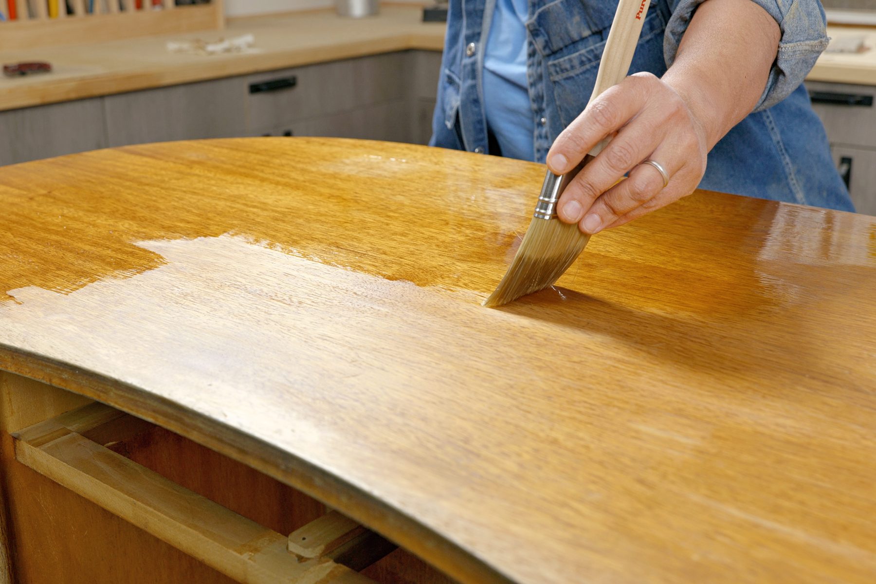 a person is applying polish on the surface of the table from a brush; wooden desk with a curved top; The wood appears to be a light-colored hardwood; The desk is placed in a workshop or garage setting, with cabinets and tools visible in the background;