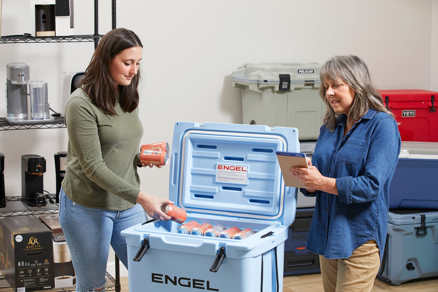 Two women are interacting with an open ENGEL cooler. One woman is placing packaged food inside the cooler, while the other is holding a tablet and taking notes. Shelves with various equipment and more coolers are visible in the background.