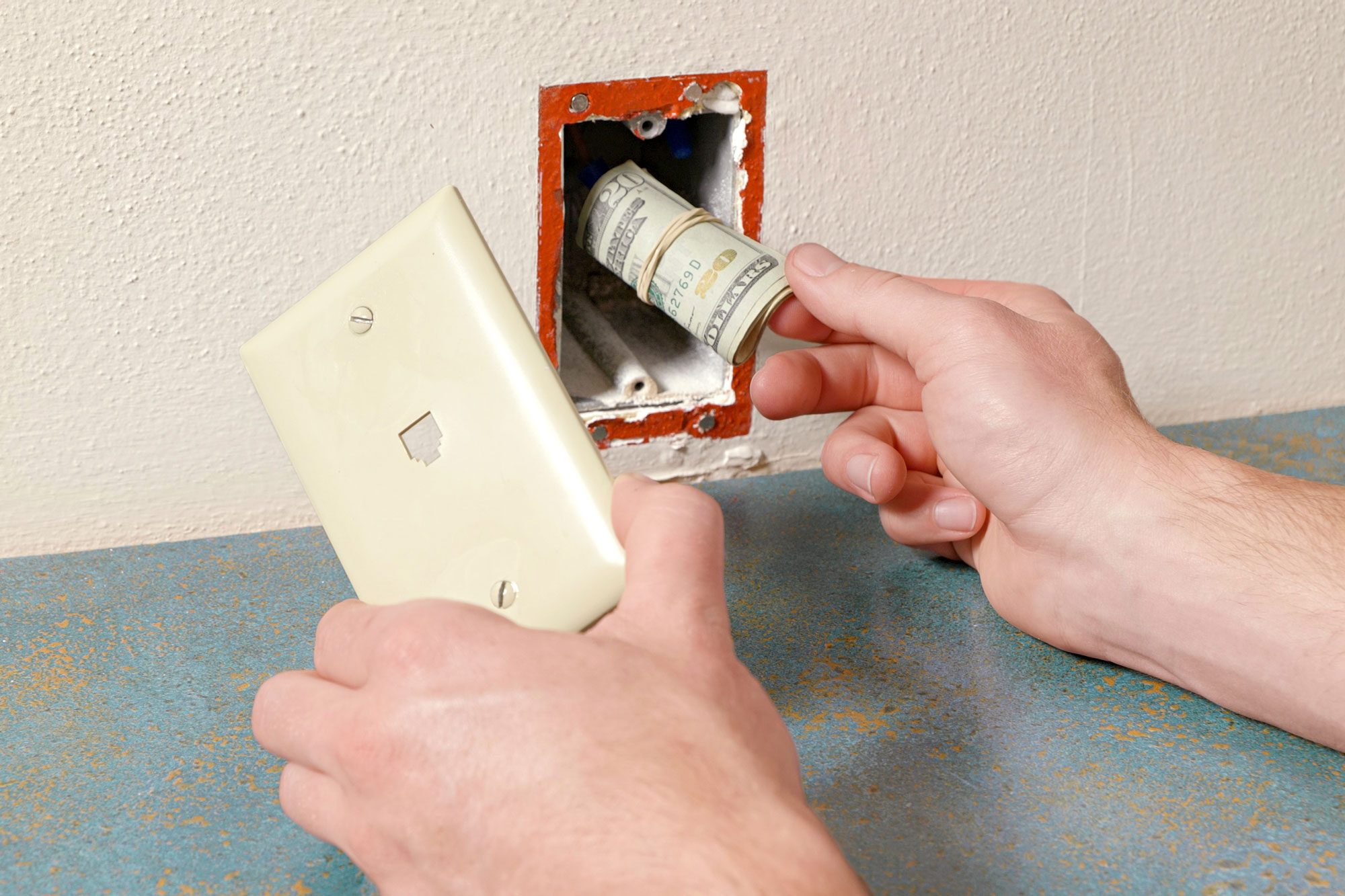 a close-up of a person's hands removing a light switch cover, Behind the switch, there’s a rolled-up banknote hidden inside the electrical box, The wall is painted in a light color, and there’s an additional electrical outlet visible to the right, The countertop appears to be made of speckled laminate in blue and brown colors;