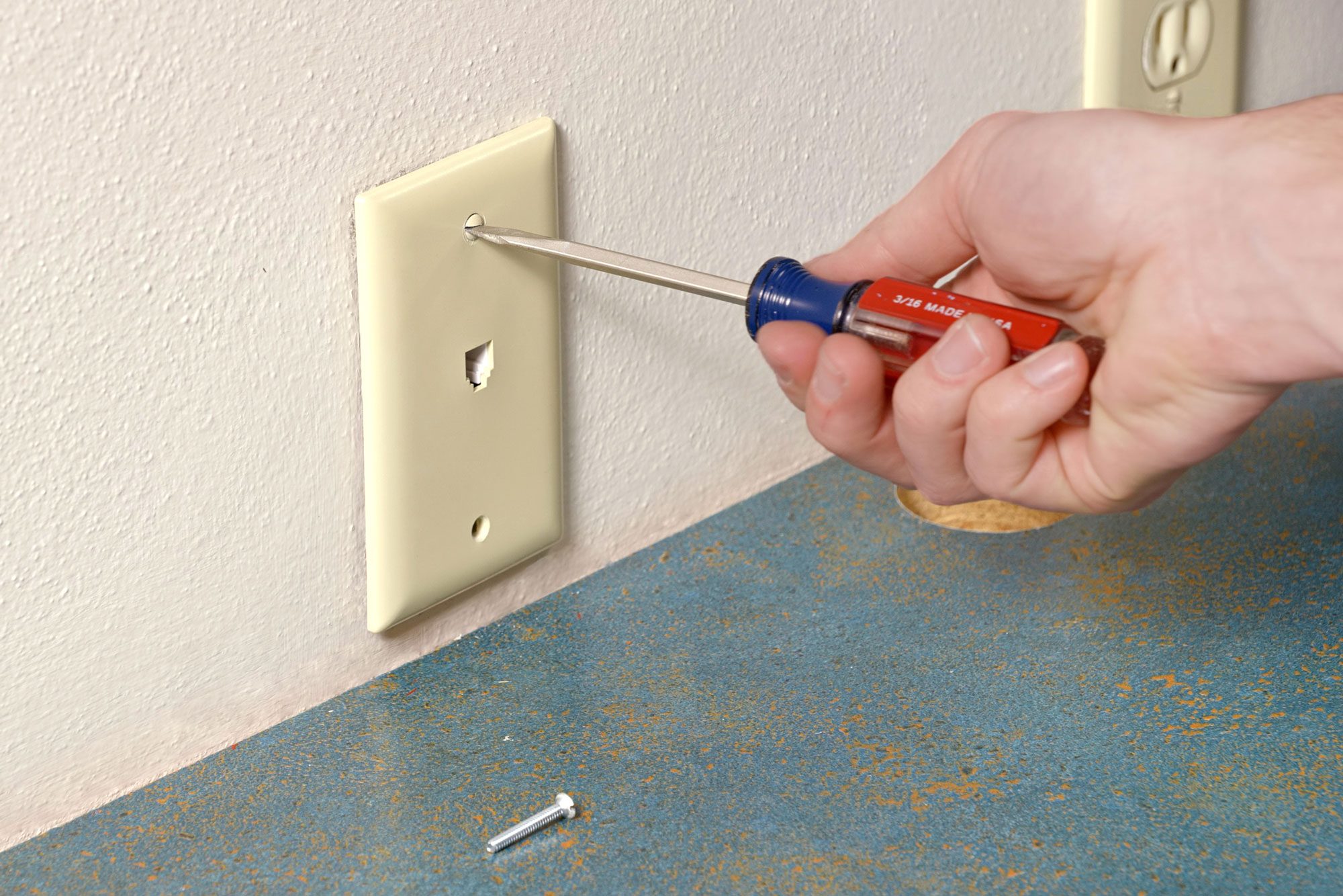 a close-up of a person’s hand using a flathead screwdriver to remove a screw from an electrical outlet cover on a wall, The wall is painted in a light color, There is one screw partially removed lying on the surface below the outlet;