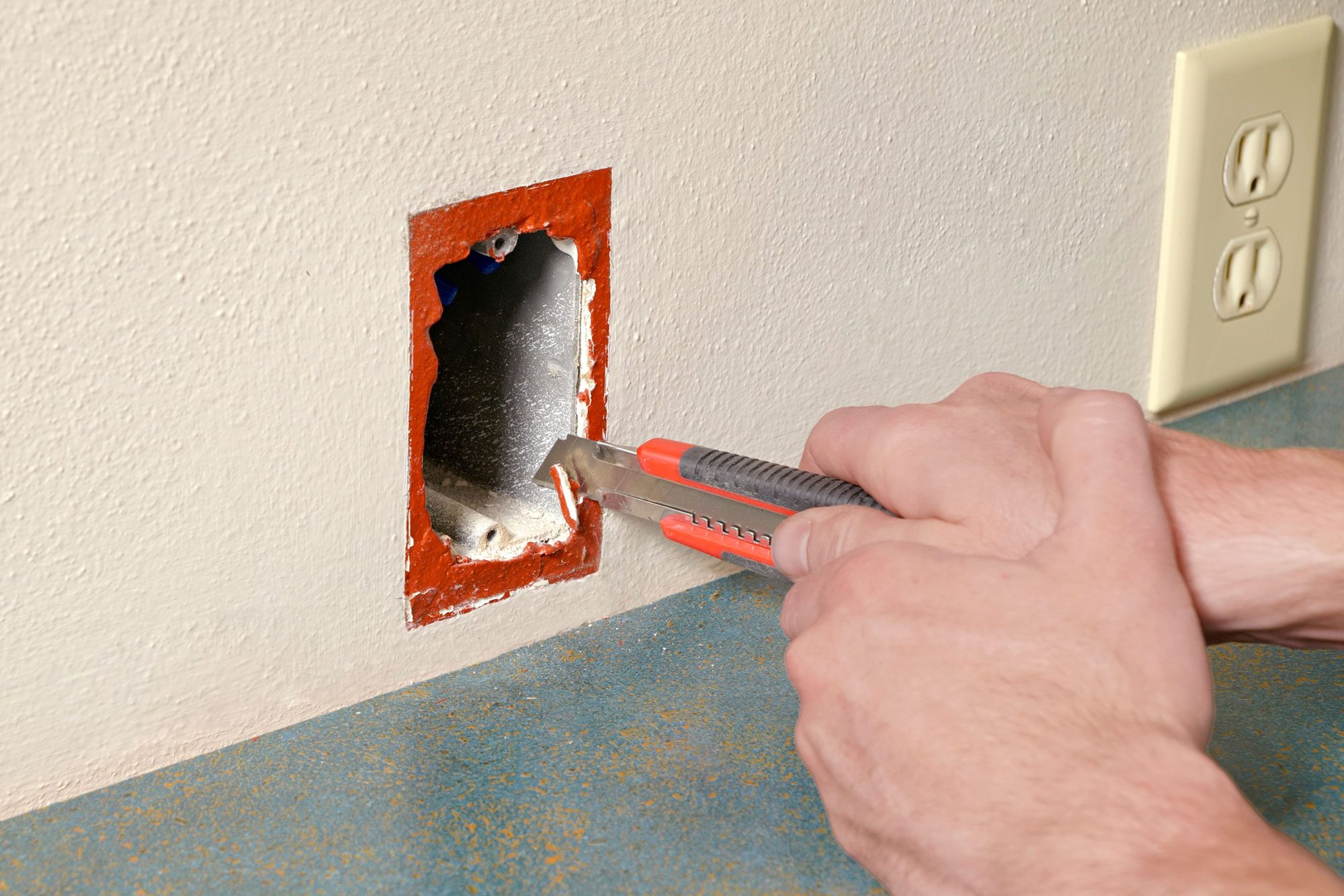 a close-up of person's hand using a red and black utility knife to cut into a section of drywall, There is a partially cut square hole in the wall, revealing some grey material behind it, To the right, there is an electrical outlet on the same wall. The wall is painted light beige;