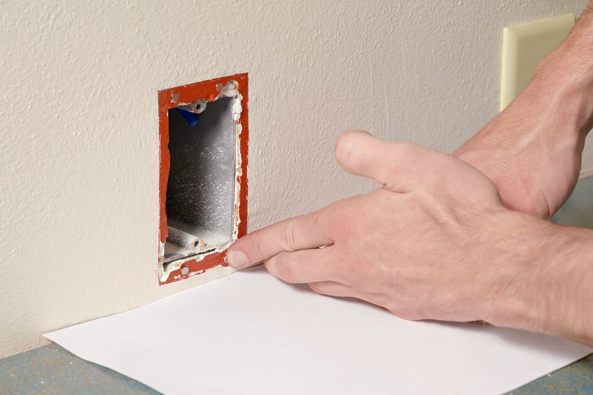 a close-up of a person’s hand pointing to an electrical box that is recessed into a wall, The wall appears to be made of drywall, and the electrical box has visible metal components inside, There is a red-orange outline around the electrical box, possibly a marker or paint, Below the wall and electrical box, there is a countertop with speckled patterns, and on top of this countertop lies a white sheet of paper;