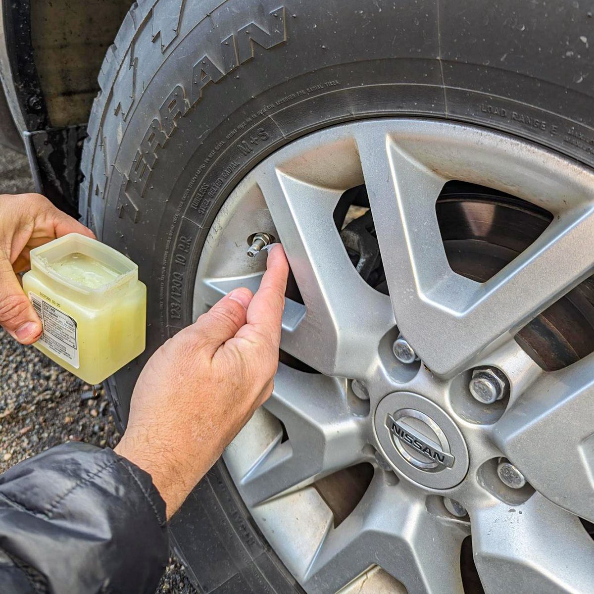 Close-up of a person applying a substance from a container to a screw on a car wheel, which has a visible "Terrain" tire and a vehicle logo in the center. The person is wearing a black jacket.