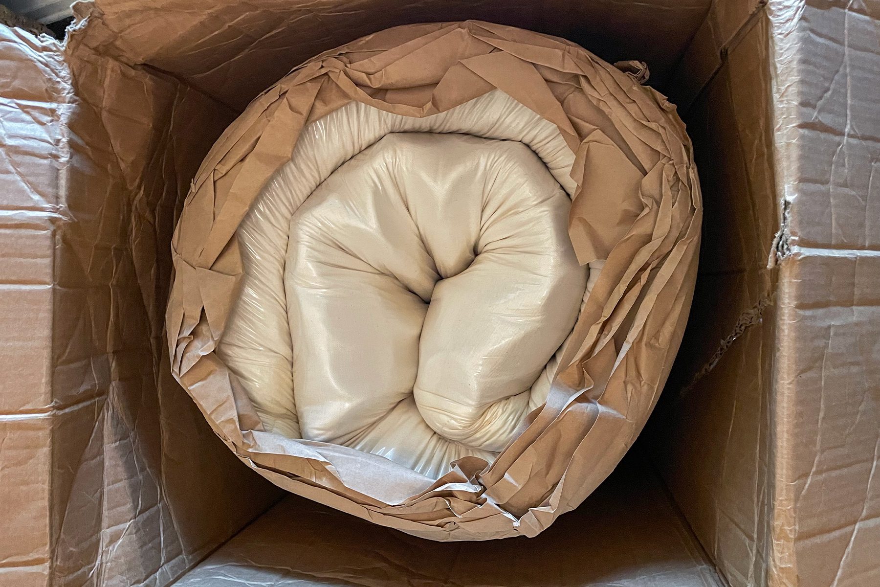 A rolled-up mattress wrapped in white fabric and brown paper is placed inside a cardboard box.