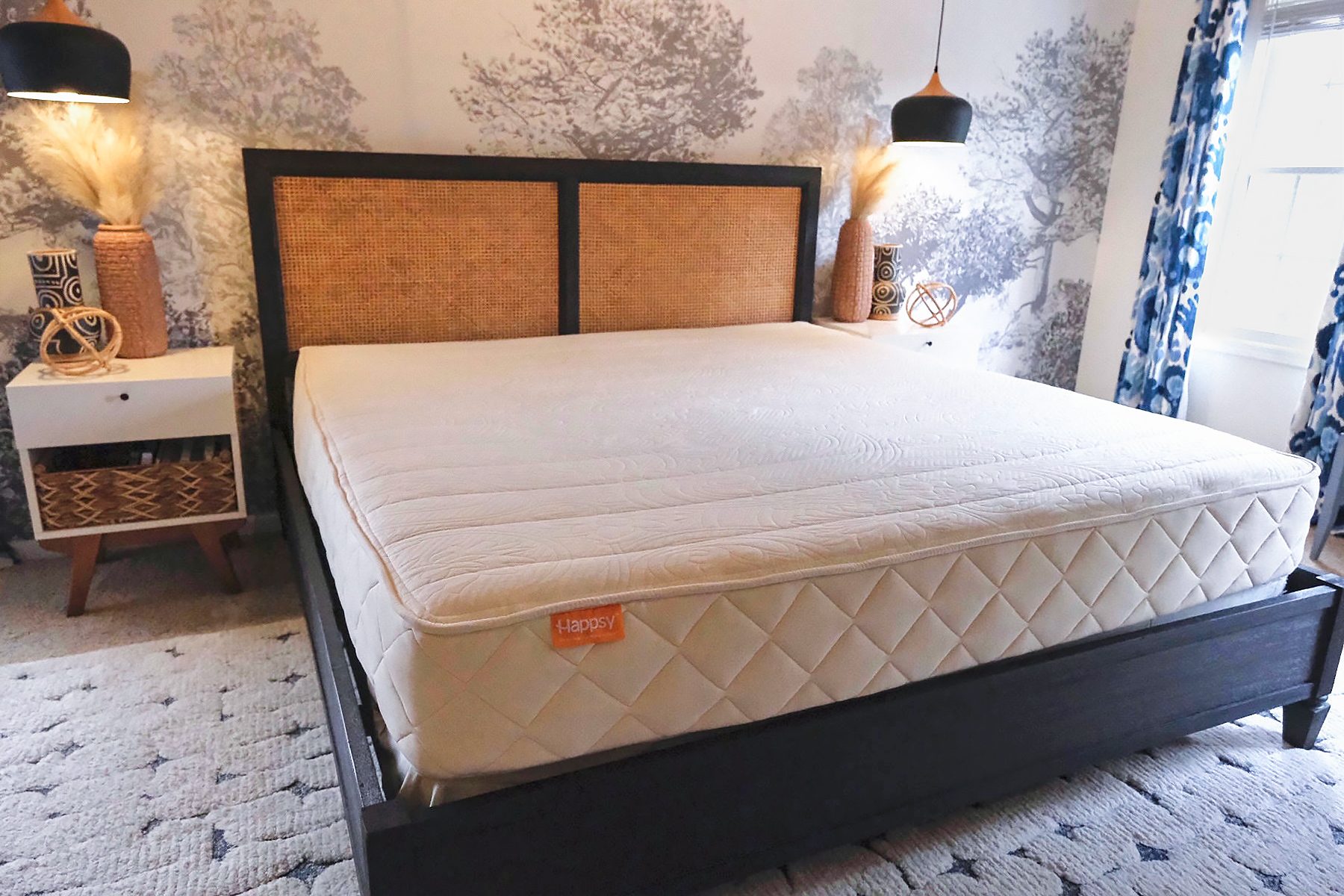 A bedroom features a large bed with a light-colored mattress and a dark wood frame. There are tree-patterned wallpaper, two hanging pendant lights, and a white nightstand with wicker baskets on the left. Blue patterned curtains adorn the window on the right.