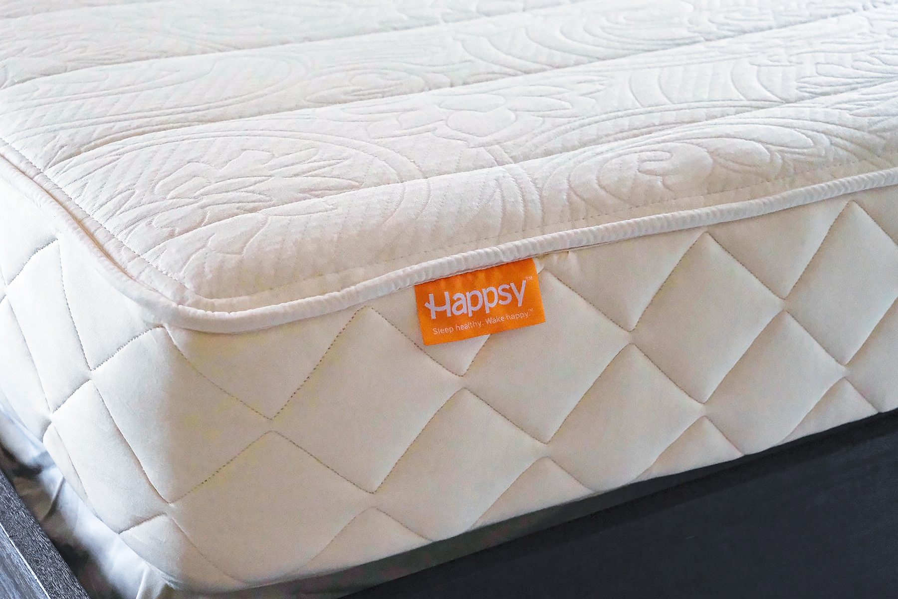 Close-up of a cream-colored, quilted mattress with an orange "Happsy" label on the side. The mattress features a textured, circular pattern on the surface and rests on a dark base.