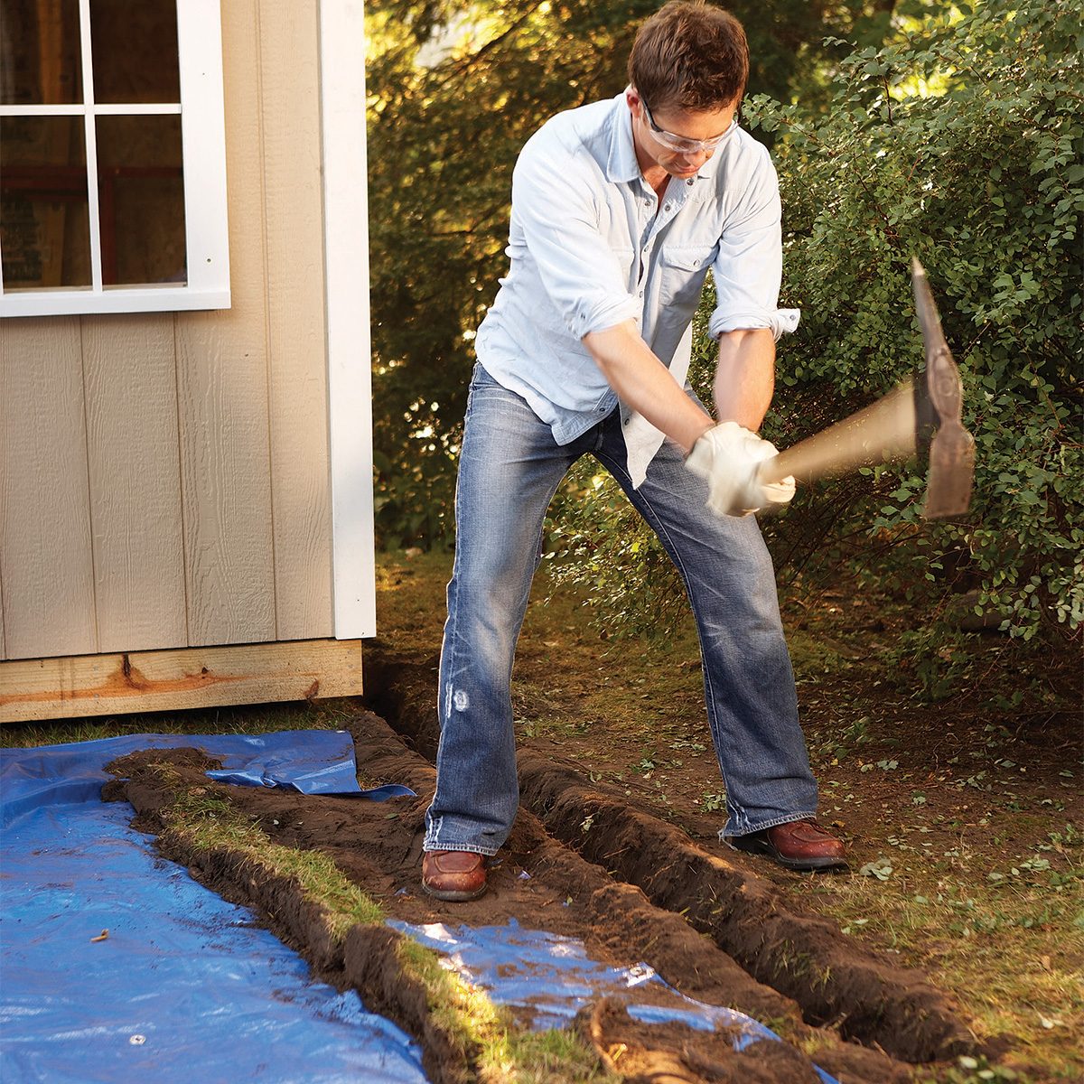 A person in blue jeans and a light shirt uses a pickaxe to dig the ground next to a wooden shed. A blue tarp is spread on the ground nearby, and there are trees in the background.