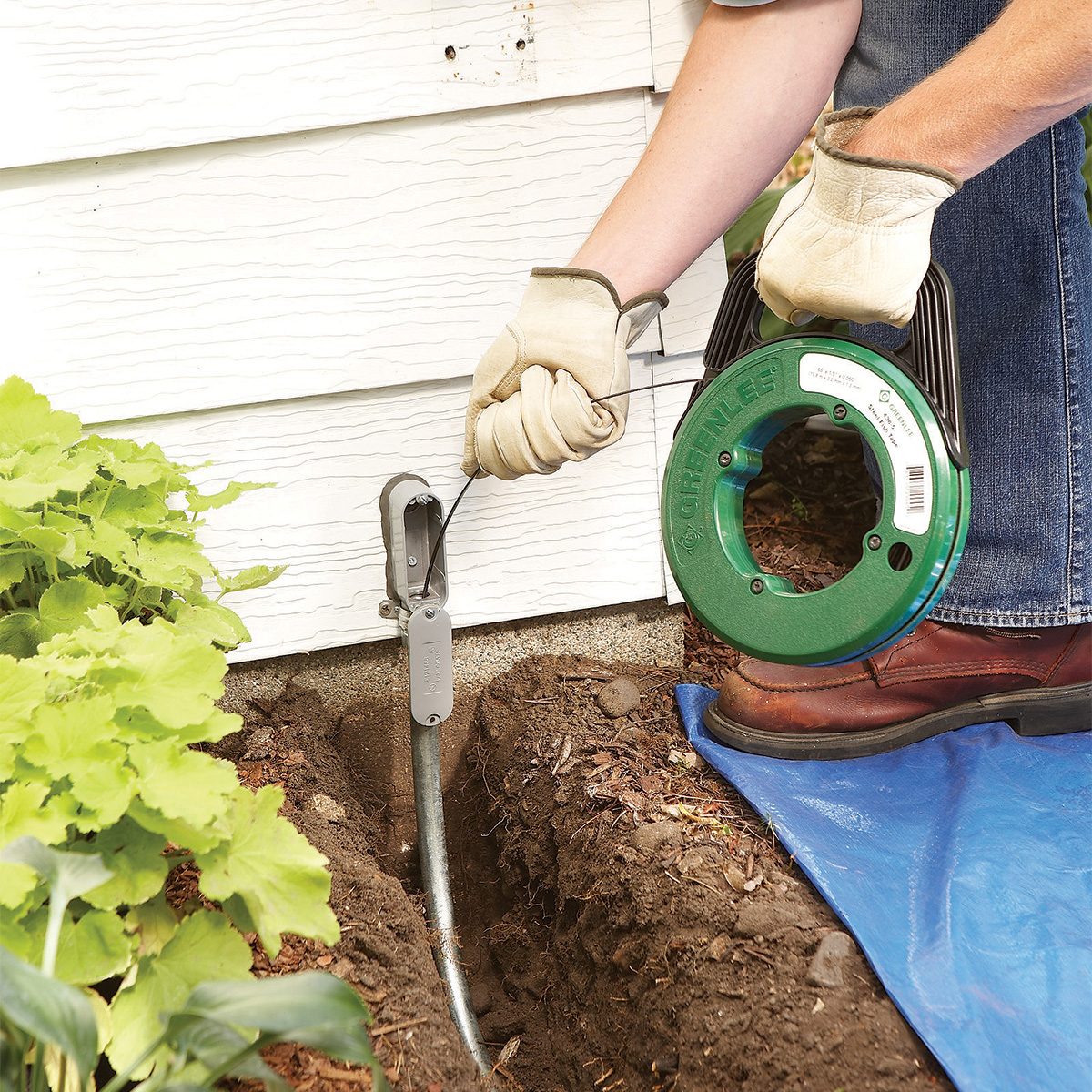 A person wearing gloves uses a green fish tape to guide wiring through a metal conduit next to a house. They are kneeling on a blue tarp, with green plants visible nearby.