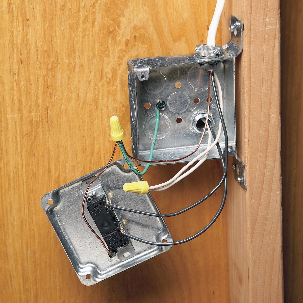 An open electrical junction box mounted on a wooden surface, showing exposed wiring with wire connectors. The metal cover is hanging to the side, revealing black, white, and green wires connected inside.