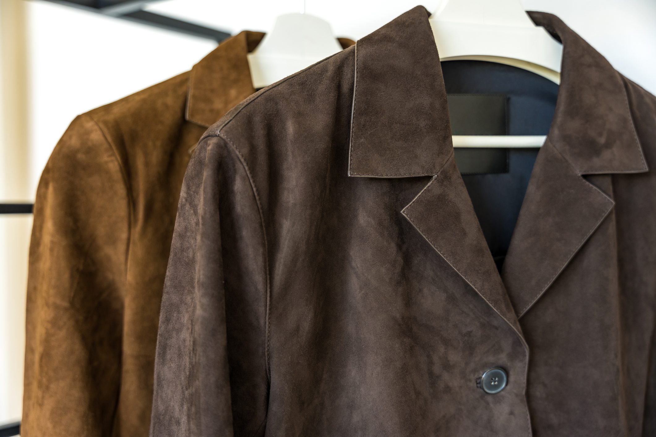 Two brown natural suede jackets of different shades hanging on a hanger on a white background.