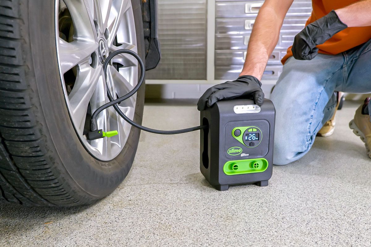 A person is inflating a car tire with a portable tire inflator; the person is wearing black gloves and jeans; they are kneeling down next to the car's tire with the inflator in hand; the inflator is plugged into the tire valve