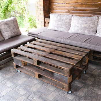 Wooden pallet coffee table on wheels in a cozy outdoor seating area with cushioned sofas. The table and sofas are placed on a tiled patio, surrounded by a brick wall and greenery in the background.